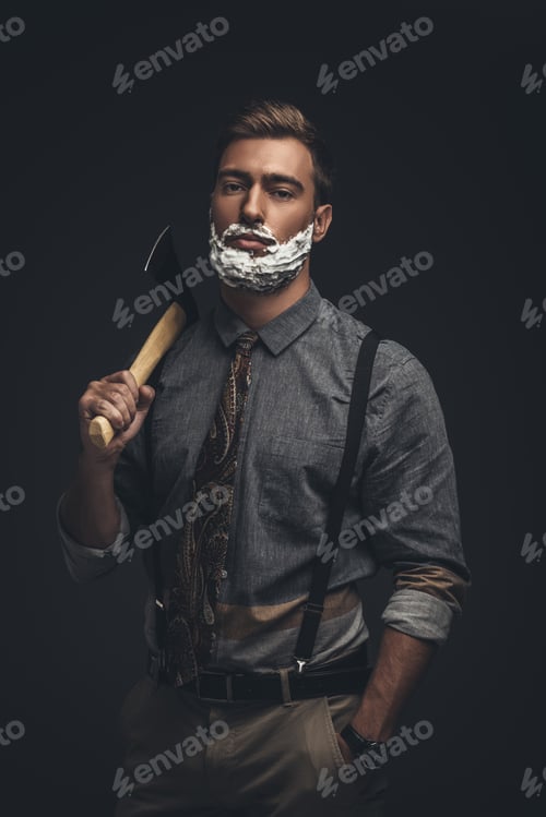 Preview: Young man with shaving cream on his face, holding an axe and looking at camera