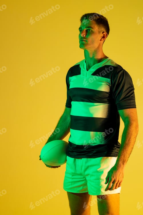 Preview: Caucasian male rugby player with rugby ball over yellow lighting