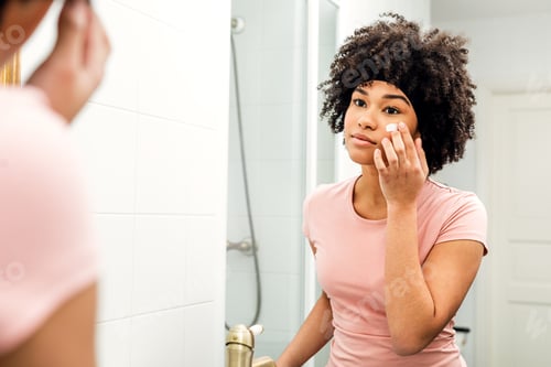 Preview: Young woman in bathroom looking in to the mirror and applying cream