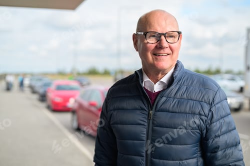 Preview: Happy senior Scandinavian man standing outdoors in front of a car park at an airport taxi area