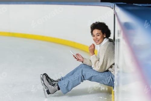 Preview: Positive african american woman holding smartphone and looking at camera on ice rink
