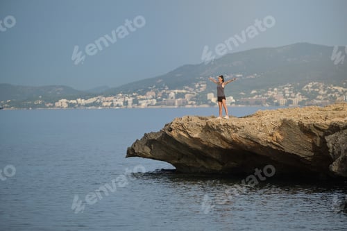 Preview: Young woman standing on stone cliff with spread arms