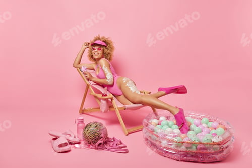 Preview: Horizontal shot of curly haired woman feels relaxed dressed in swimwear applies sunscreen lotion