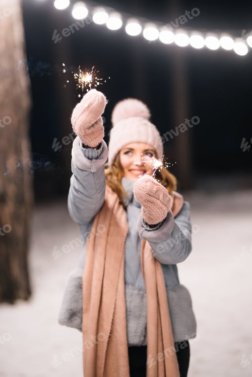 Preview: Beautiful girl with sparklers in hands. Happy winter time in the forest. Festive garland lights.