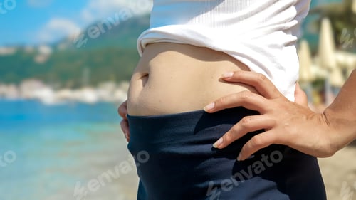 Preview: Closeup of young woman with excess fat on belly standing on the sea beach during summer holiday