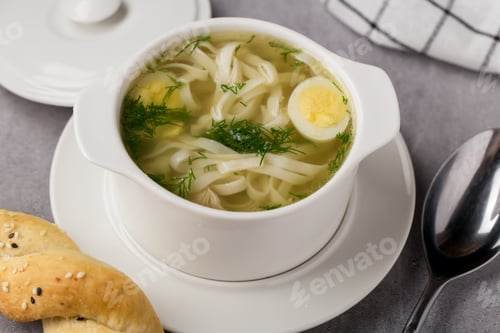 Preview: Hot soup with chicken, noodles and egg in a white bowl, with a bun