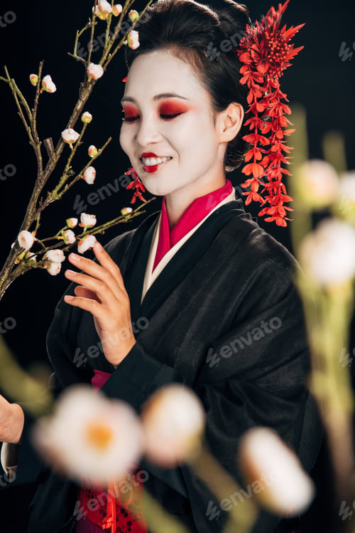 Preview: selective focus of happy beautiful geisha in black kimono with red flowers in hair and sakura