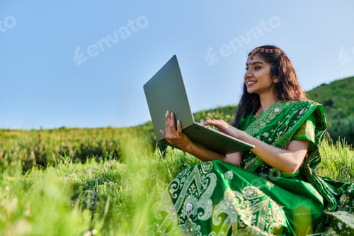 Preview: smiling young indian woman in green sari using laptop on grassy meadow in summer