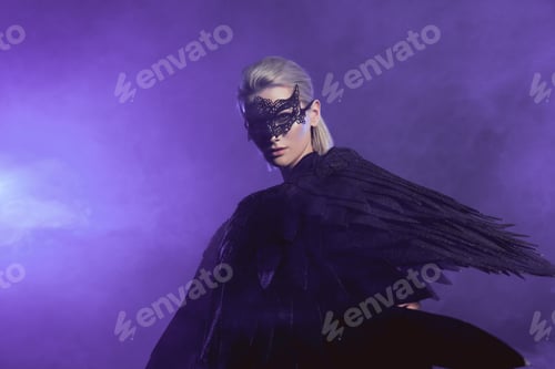 Preview: beautiful woman in lace mask and black angel wings posing on smoky violet background with copy space