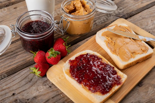 Preview: Close-up peanut butter and jelly sandwich on serving board with jars, strawberries and milk at table