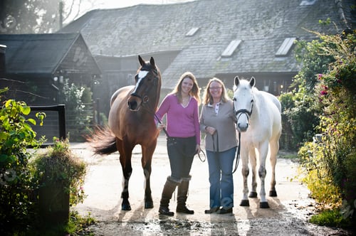 Preview: Two women standing outside a stable with two horses.
