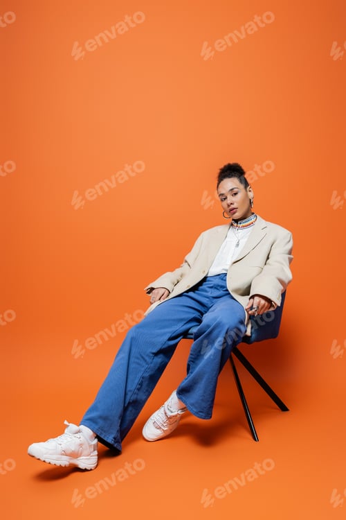 Preview: beautiful african american fashion model in street vibrant outfit sitting relaxed on blue chair