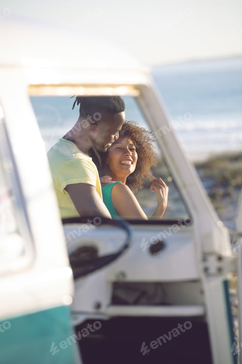 Preview: Side view of young African american man embracing woman near camper van at beach
