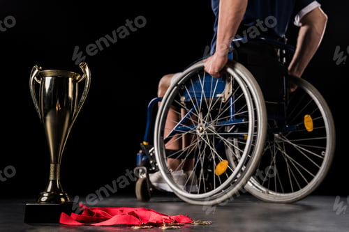 Preview: cropped view of paralympic in wheelchair going away from his champion goblet and gold medals