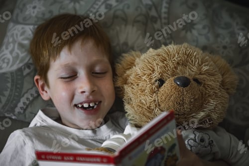 Preview: Child 6-7 years old red-haired boy lies in bed reading a book before going to bed with a teddy bear