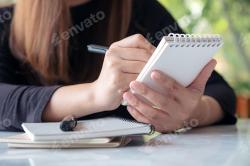 Preview: A woman holding and writing on notebook while sitting in cafe with blur green nature background