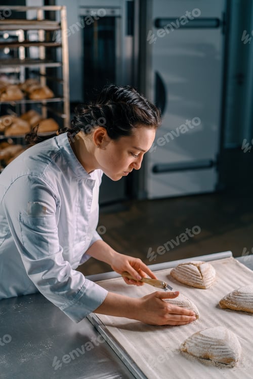 Preview: beautiful woman baker cuts patterns with a professional baker's knife on raw bread buns