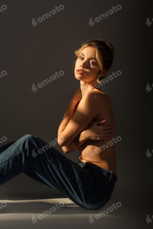 Preview: pretty half nude woman in jeans sitting with crossed arms and looking at camera on grey background