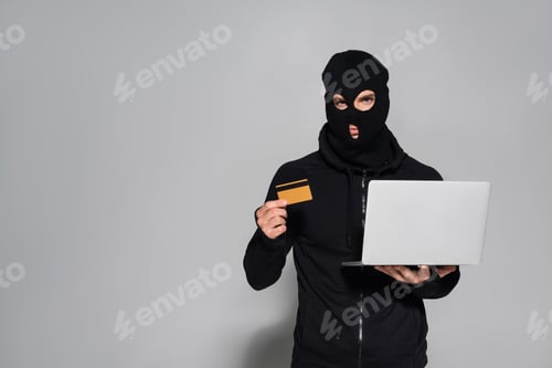 Preview: Hacker in balaclava holding credit card and laptop on grey background