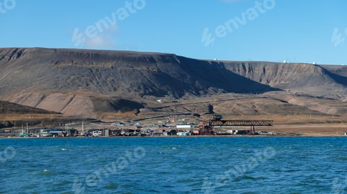 Preview: A coastal settlement in Svalbard is set against towering mountains with clear blue skies above
