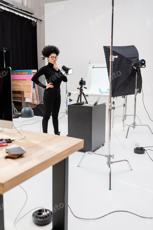 Preview: pensive african american content maker standing with hand on hip near laptop and softbox reflector