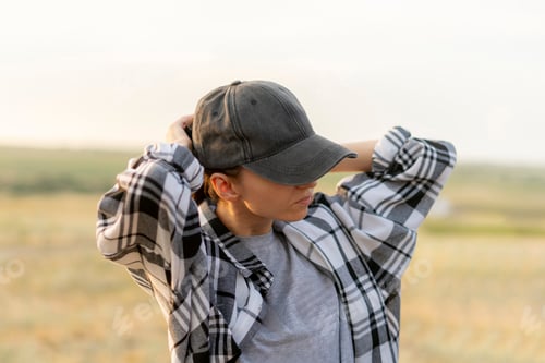 Preview: a woman wearing a plaid shirt and stands in a field. She puts on a black cap and adjusting it