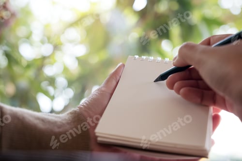 Preview: Closeup image of hands holding and writing down on a blank notebook with blur nature background