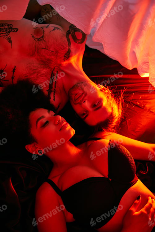 Preview: attractive multicultural couple lying sensually together under bedsheet surrounded by lights