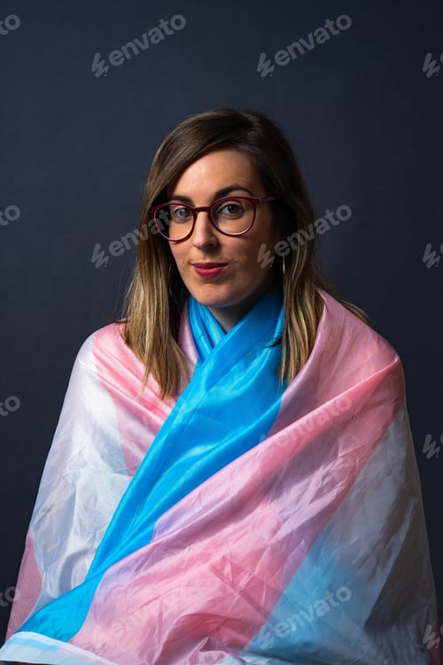 Preview: Blonde Transgender Woman with Glasses and LGTB Trans Flag Cape looking at camera in Studio