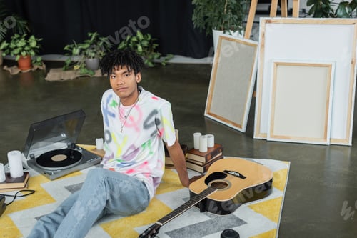 Preview: African american artist looking at camera near acoustic guitar and canvases in workshop