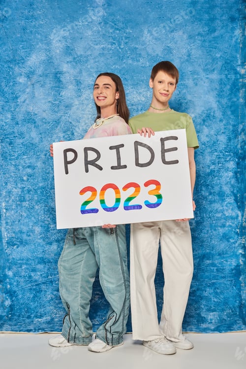 Preview: Full length of cheerful queer friends looking at camera while holding placard with pride 2023