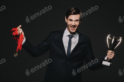 Preview: happy businessman wearing medals and celebrating victory while holding trophy isolated on black
