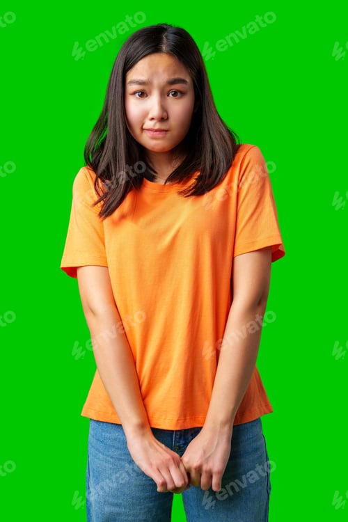 Preview: Girl in orange shirt expressing shyness against a bright green background indoors