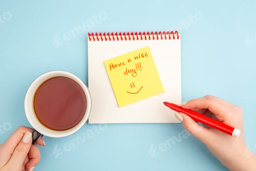 Preview: top view spiral notebook cup of tea and red marker in female hands have a nice day written on sticky