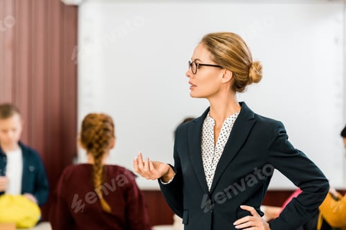 Preview: young female teacher in eyeglasses looking away while schoolkids standing behind