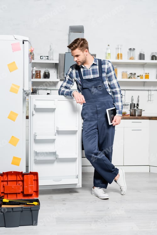 Preview: full length of young handyman with tablet looking at fridge in kitchen
