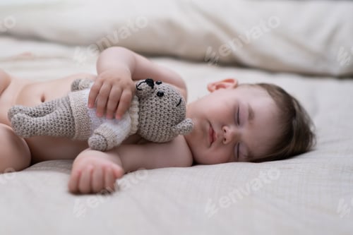 Preview: Innocent baby boy with closed eyes sleeping on soft bed with white sheets hugging knitted animal toy