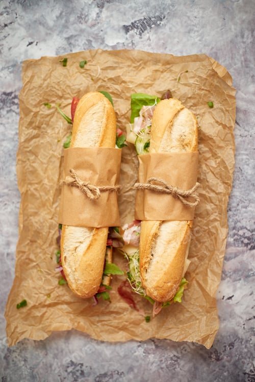 Preview: Sandwiches with ham, fresh vegetables and herbs served on brown baking paper over concrete backdrop