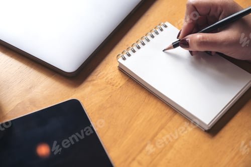 Preview: woman's hand writing on a blank notebook with laptop , tablet and coffee cup on wooden table
