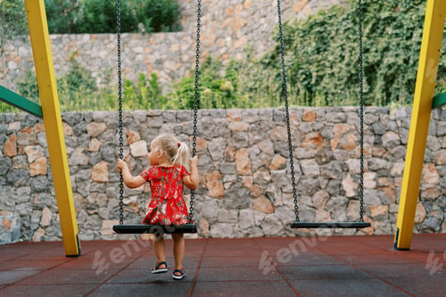 Preview: Little girl sits on a chain swing on a playground in the yard and looks away. Back view