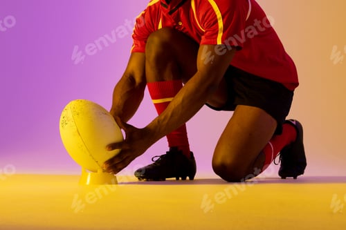 Preview: African american male rugby player crouching with rugby ball over pink lighting
