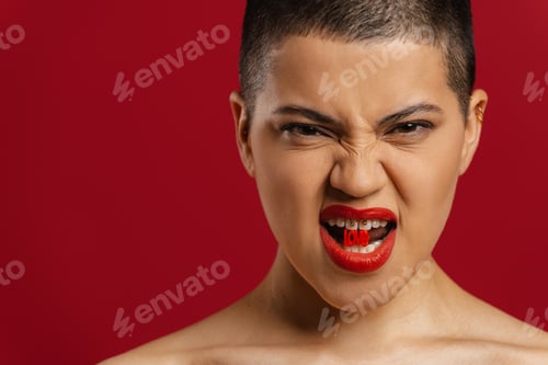 Preview: Young shaved head woman holding plastic letters in mouth and grimacing against red background