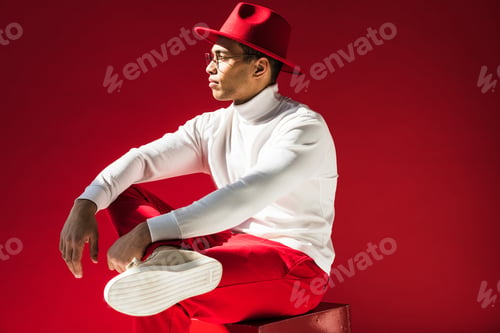 Preview: side view of stylish mixed race man in hat and glasses sitting and posing isolated on red
