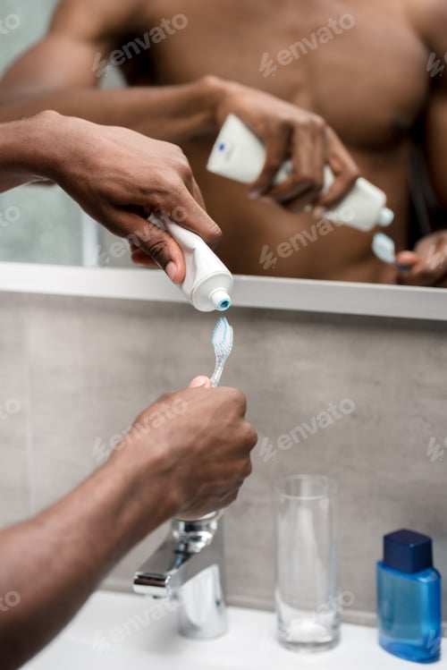 Preview: cropped shot of young african american man holding toothpaste and toothbrush in bathroom