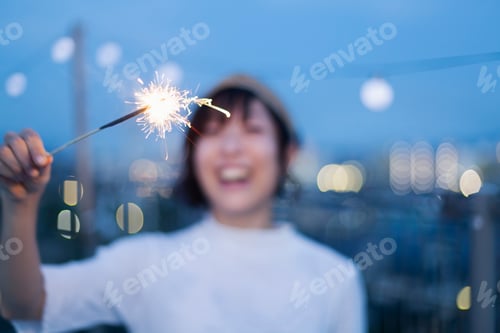 Preview: Smiling young Japanese woman holding sparkler on a rooftop in an urban setting.