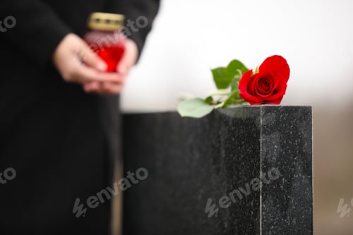 Preview: Woman with candle near black granite tombstone outdoors, focus on red rose. Funeral ceremony