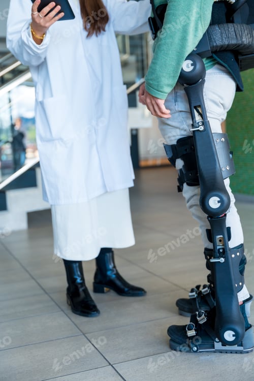 Preview: Female physiotherapist doctor talking smiling with tablet with disabled person with robotic skeleton