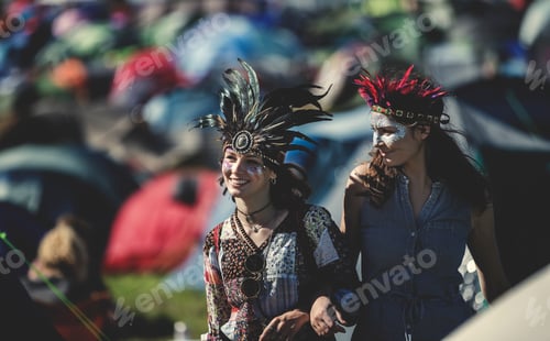 Preview: Two smiling young women at a summer music festival face painted, wearing feather headdress, standing
