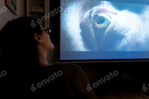 Preview: Young woman, girl with glasses watches tv, movie at night on huge screen at home on couch in room. I