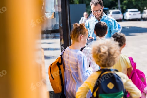 Preview: rear view of pupils entering school bus while teacher writing in clipboard